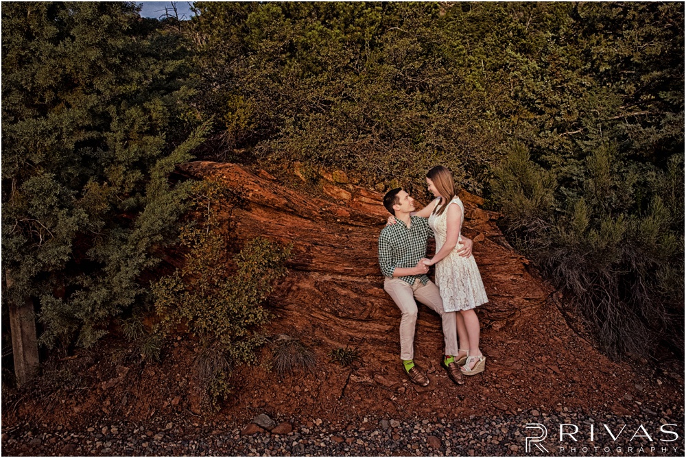 Merry-Go-Round Rock Engagement Session | Picture of an engaged couple sitting in front of red rocks and green shrubs at Merry-Go-Round Rock in Sedona.  