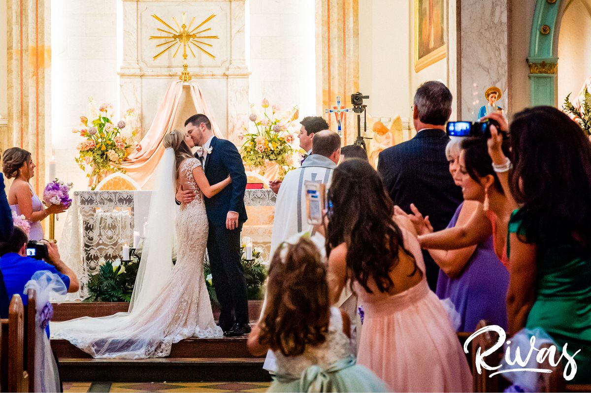 8 Things Your Wedding Photographer Wants you to Know | A photo of a bride and groom sharing their first kiss while half of the audience leans out in the aisle to take photos on their phone. 