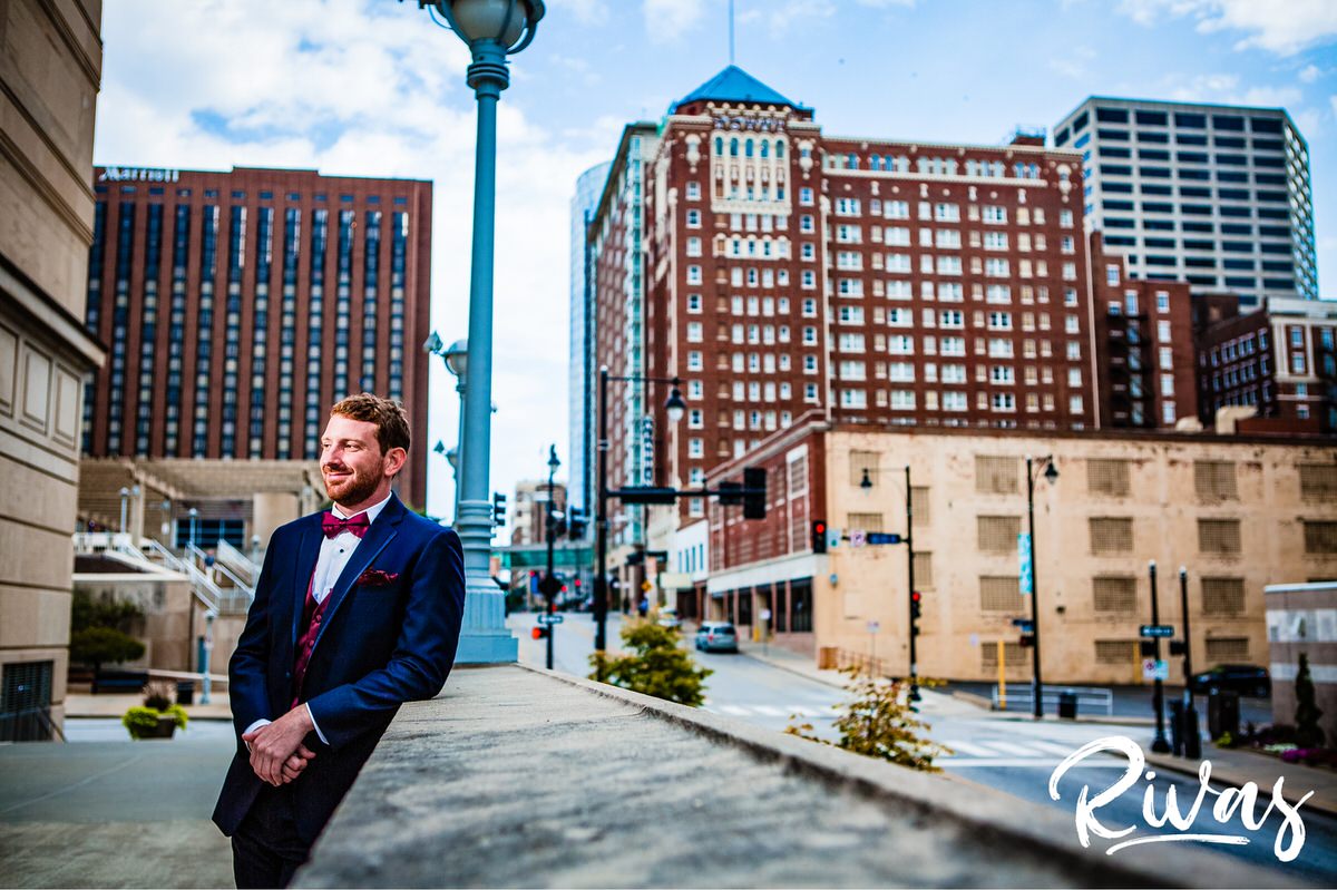 Strawberry Hill Summer Wedding | Rivas Photography | Kansas City Wedding Pictures | A relaxed picture of a groom leaning up against a cement wall and smiling while looking away from the camera in front of downtown Kansas City's Barney Allis Plaza on the morning of his wedding. 