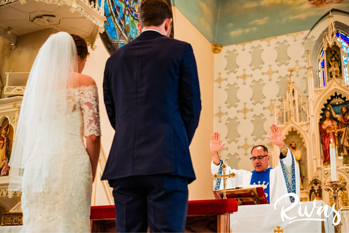 Strawberry Hill Summer Wedding | Rivas Photography | Kansas City Wedding Pictures | wide picture taken from the front of the church of a bride and groom standing together as the priest raises his hands to give the final blessing to the couple on their wedding day. 