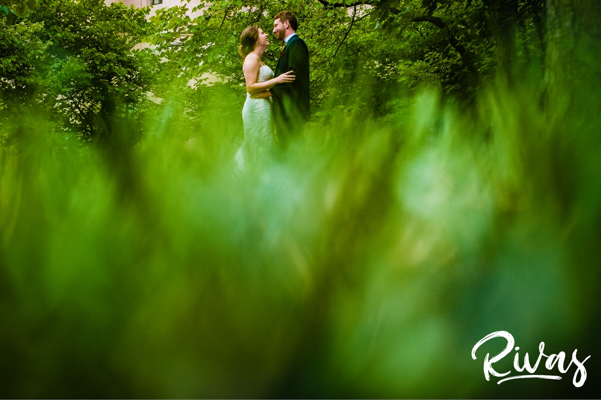 Strawberry Hill Summer Wedding | Rivas Photography | Kansas City Wedding Pictures | A vibrant photo taken through the grass looking up as a bride and groom share an embrace and laugh together while standing outside Kansas City's Nelson Atkins Museum of Art on their wedding day. 