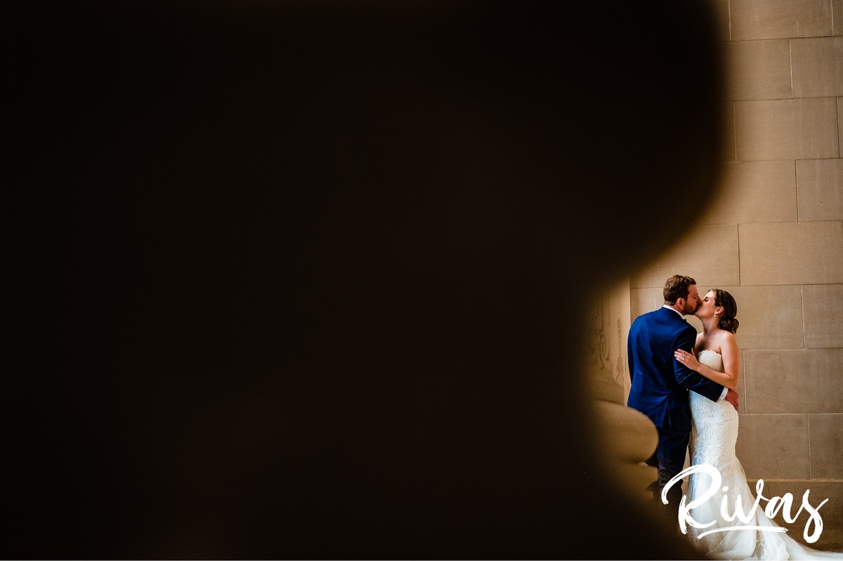 Strawberry Hill Summer Wedding | Rivas Photography | Kansas City Wedding Pictures | A dramatic photo of a bride and groom embracing and sharing a kiss on their wedding day, taken through the curve of the base of a column at Kansas City's Nelson Atkins Museum of Art. 