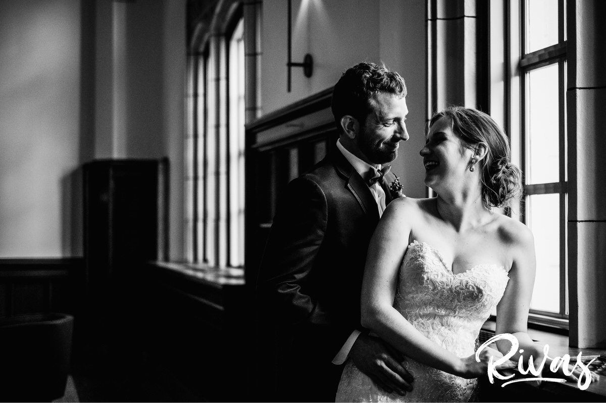 Strawberry Hill Summer Wedding | Rivas Photography | Kansas City Wedding Pictures | A black and white, candid photo of a bride looking over her shoulder and laughing at her groom as they stand in front of the windows on the main level of The Brass on Baltimore. 