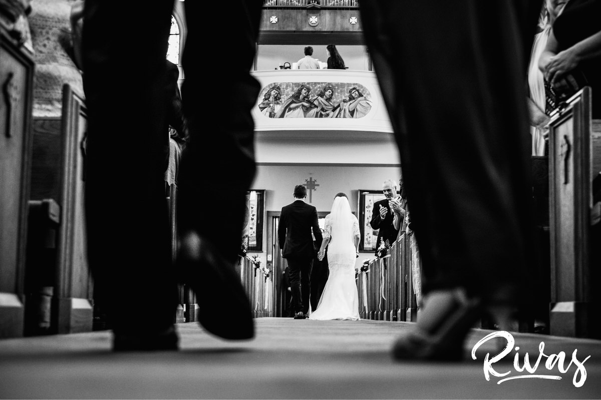 Strawberry Hill Summer Wedding | Rivas Photography | Kansas City Wedding Pictures | A black and white picture taken from the ground up and in between a pair of legs walking towards the back of the church. The bride and groom are in focus in between the legs at the back of the church holding hands after their wedding ceremony. 