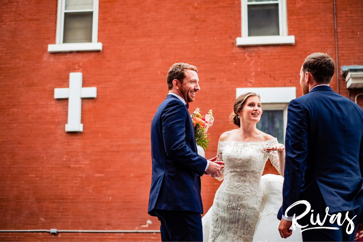 Strawberry Hill Summer Wedding | Rivas Photography | Kansas City Wedding Pictures | A candid picture of a bride and groom turning and laughing with their wedding party in front of a red brick wall with a white cross visible on their wedding day. 