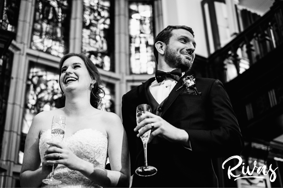 Strawberry Hill Summer Wedding | Rivas Photography | Kansas City Wedding Pictures | A black and white picture of a bride and groom holding champagne flutes and laughing as their friends toast them during their wedding reception in The Tudor Room at The Brass on Baltimore in downtown Kansas City. 