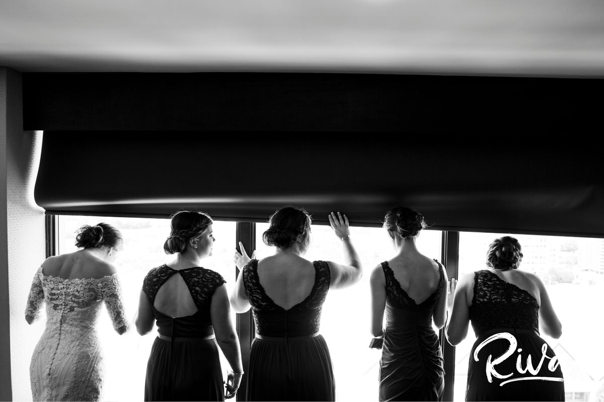 Strawberry Hill Summer Wedding | Rivas Photography | Kansas City Wedding Pictures | A candid, black and white picture of a bride and her bridesmaids looking out the window as they watch the groom and his groomsmen make their way to the church on the morning of his wedding in Kansas City. 