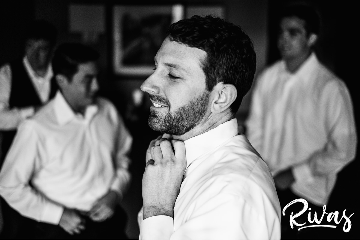 Strawberry Hill Summer Wedding | Rivas Photography | Kansas City Wedding Pictures | A close-up, black and white candid image of a groom buttoning his collar with his groomsmen visible in the background on his Kansas City wedding day. 