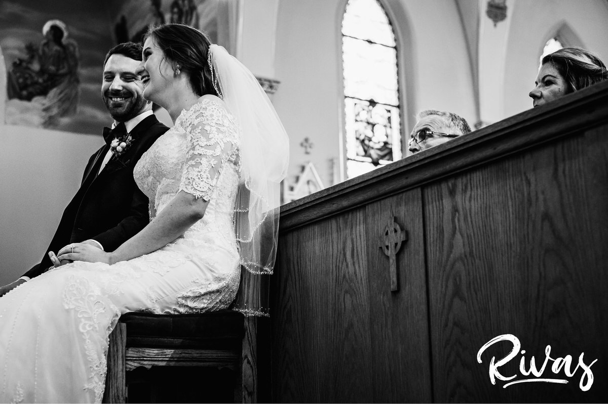 Strawberry Hill Summer Wedding | Rivas Photography | Kansas City Wedding Pictures | A black and white picture taken from the front of the church of the bride and groom sitting together laughing and holding hands during their wedding ceremony as the bride's parents sit behind them looking on. 