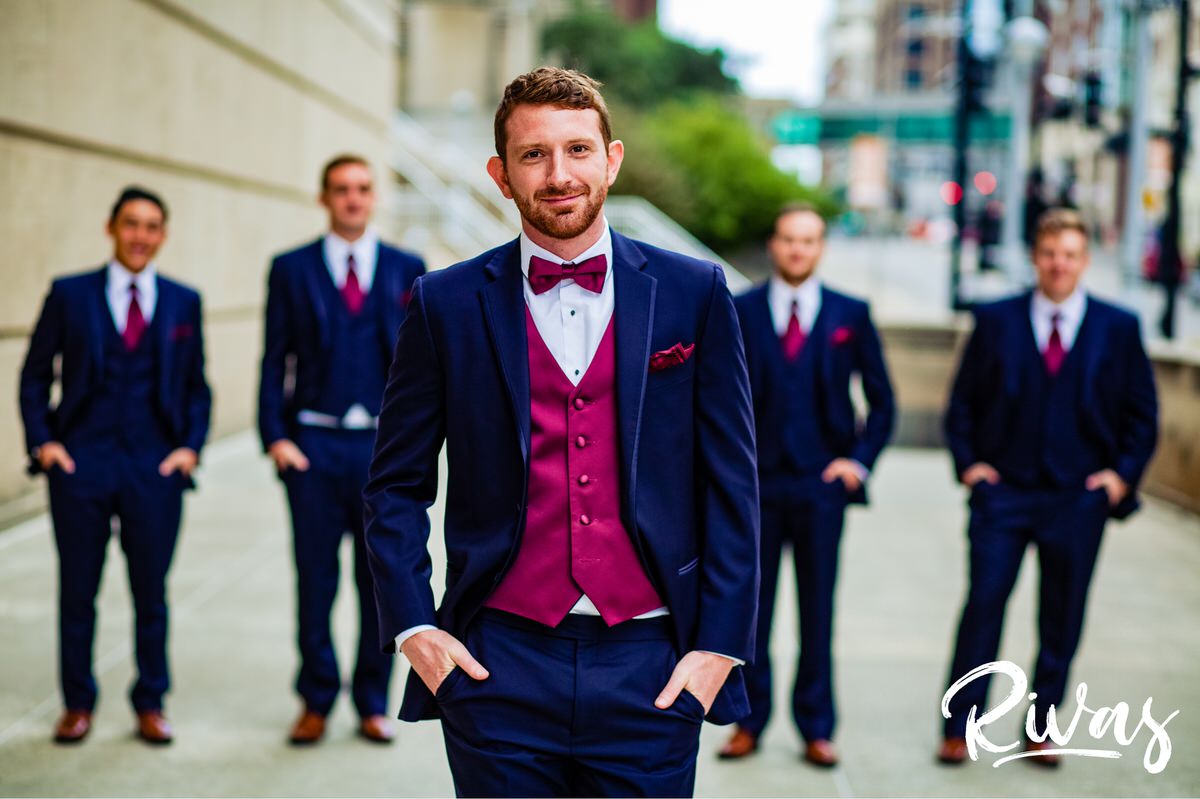 Strawberry Hill Summer Wedding | Rivas Photography | Kansas City Wedding Pictures | A candid picture of a groom walking towards the camera with his hands in his pocket with his groomsmen standing in the background at Kansas City's Barney Allis Plaza on the morning of his wedding. 