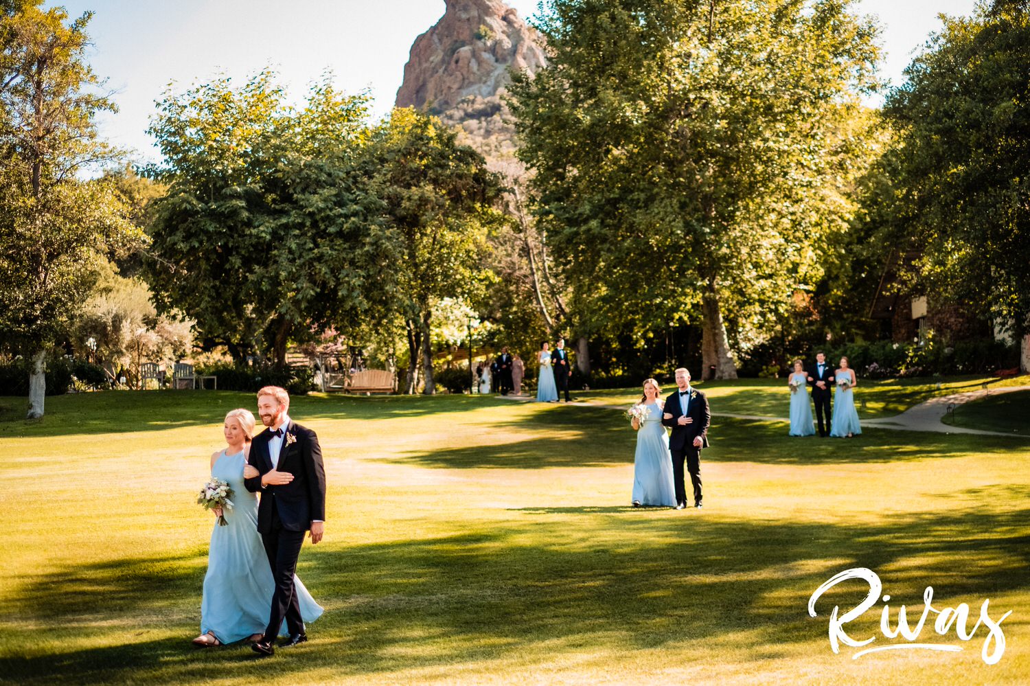Saddlerock Ranch Summer Wedding | Destination Wedding Photographers | Rivas | A wide photo of a bridal party dressed in light blue dresses and black tuxedos walking arm in arm down the lawn to the wedding ceremony cite. 