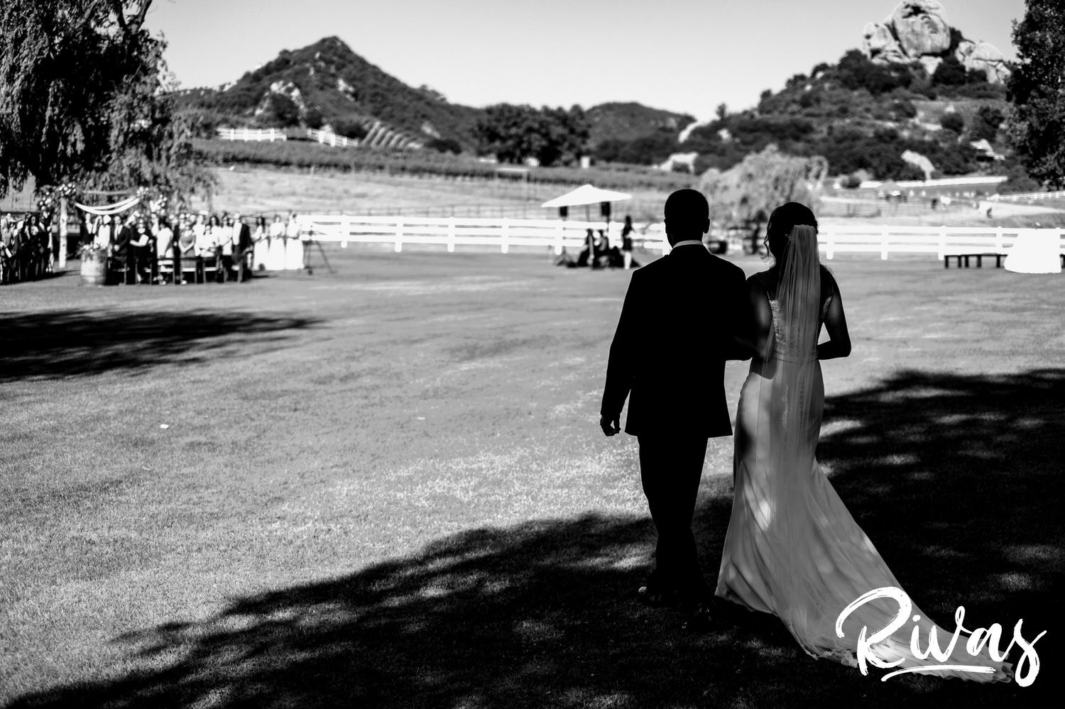 Saddlerock Ranch Summer Wedding | Destination Wedding Photographers | Rivas | A black and white image of a bride and her father walking across the lawn to the wedding ceremony at Saddlerock Ranch, taken from behind at their point of view. 