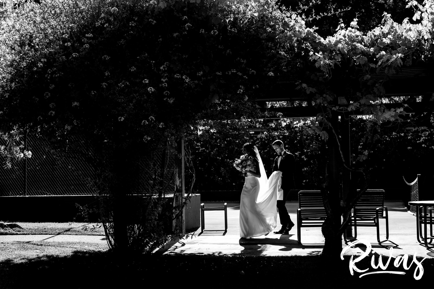 Saddlerock Ranch Summer Wedding | Destination Wedding Photographers | Rivas | A black and white image of a bride and groom walking underneath an arbor of flowers while the groom carries the bride's train and veil. 