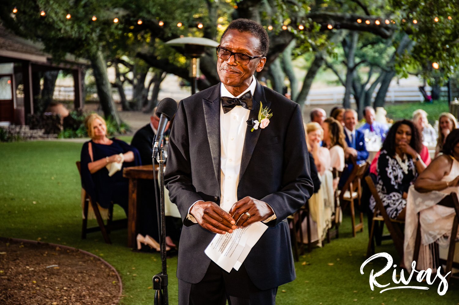 Saddlerock Ranch Summer Wedding | Destination Wedding Photographers | Rivas | A picture of a bride's dad in his tuxedo smiling at his daughter and son-in-law as he gives his toast during their wedding reception at Saddlerock Ranch in Malibu. 