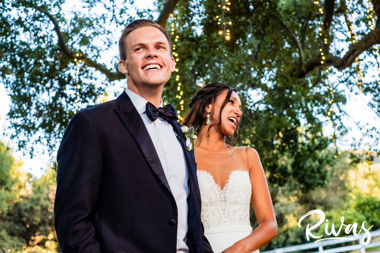 Saddlerock Ranch Summer Wedding | Destination Wedding Photographers | Rivas | A picture of a bride and groom holding hands and laughing out loud while their family toasts them during their wedding reception at Malibu's Saddlerock Ranch. 