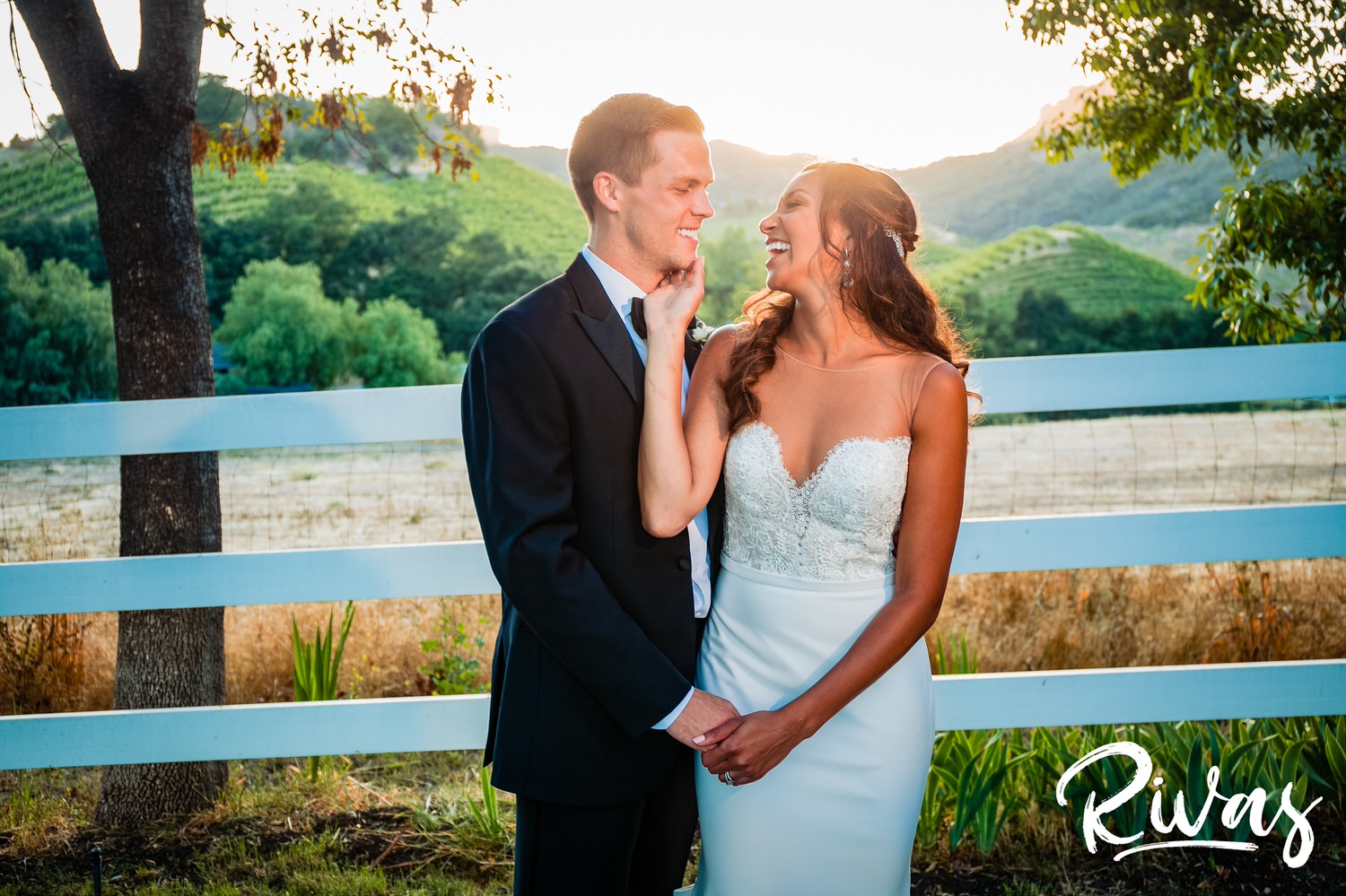 Saddlerock Ranch Summer Wedding | Destination Wedding Photographers | Rivas | A candid picture of a bride touching her groom's face with a loving touch while standing in front of a white fence line at Saddlerock Ranch in Malibu. 