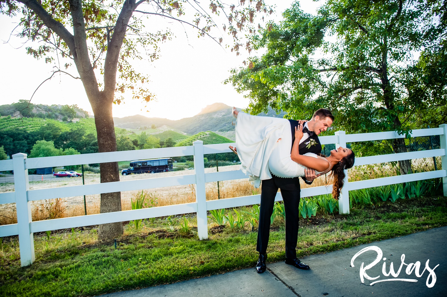 Saddlerock Ranch Summer Wedding | Destination Wedding Photographers | Rivas | A candid picture of a groom dipping his bride backwards while standing in front of a white fence line at Saddlerock Ranch in Malibu. 