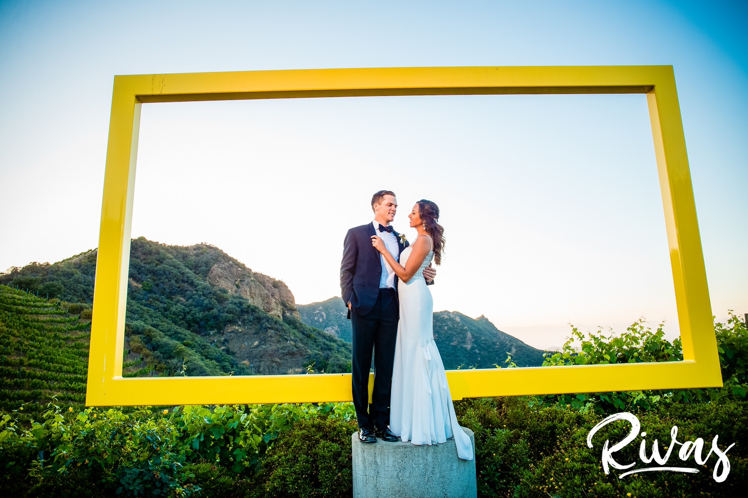 Saddlerock Ranch Summer Wedding | Destination Wedding Photographers | Rivas | A photo of a bride and groom sharing an embrace on their wedding day in front of the iconic yellow frame at Saddlerock Ranch in Malibu, California. 