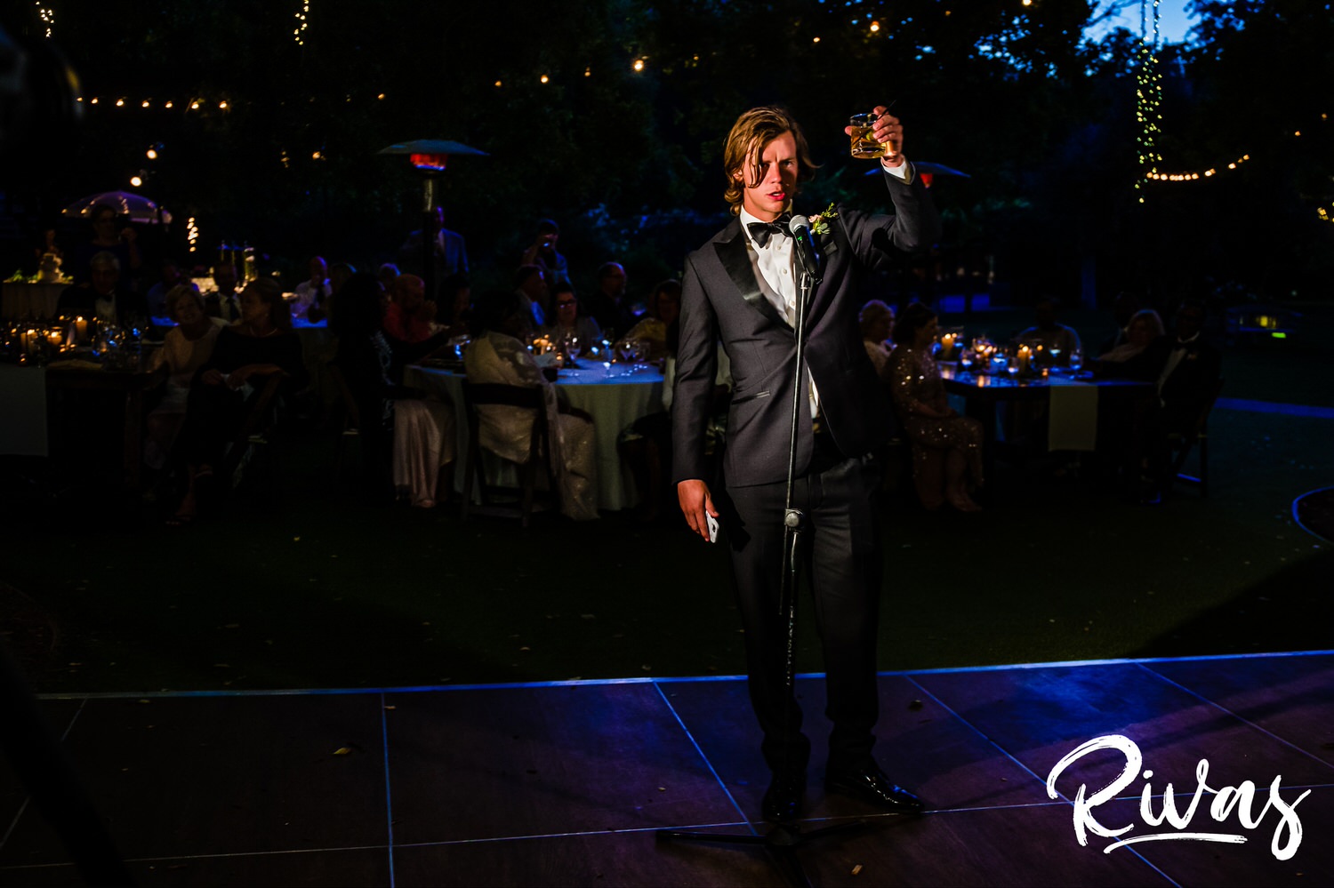 Saddlerock Ranch Summer Wedding | Destination Wedding Photographers | Rivas | A photo of a best man in a black tuxedo raising his glass as he gives a toast to the bride and groom during their Saddlerock Ranch wedding reception. 