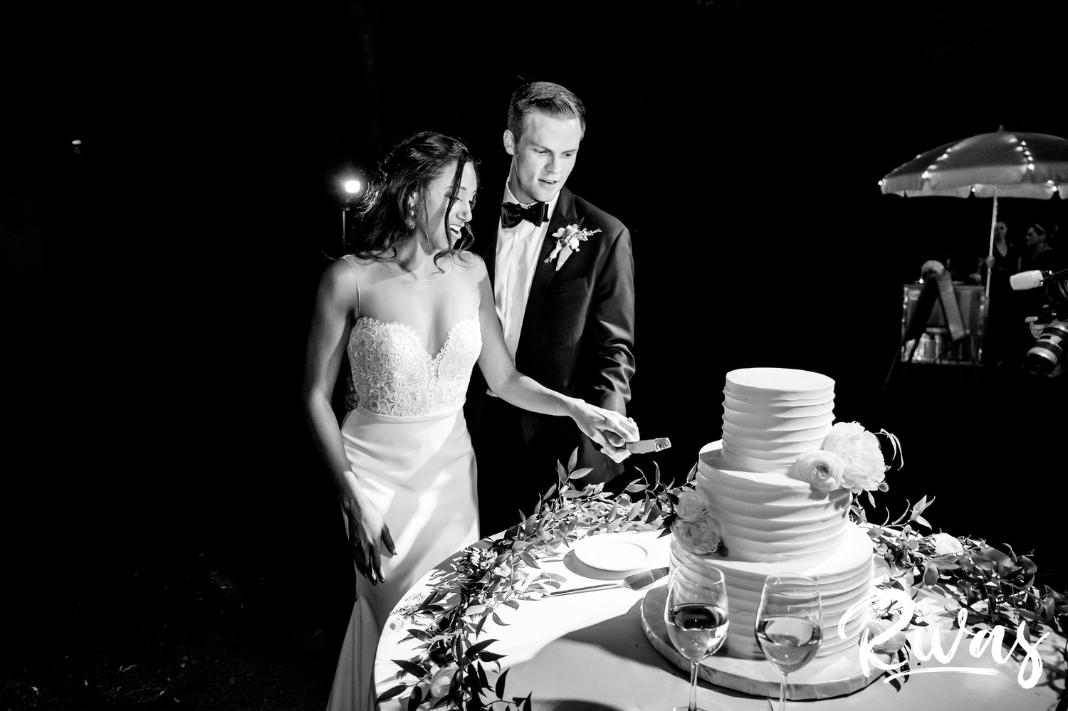 Saddlerock Ranch Summer Wedding | Destination Wedding Photographers | Rivas | A black and white candid image of a bride and groom cutting their three tier, white buttercream wedding cake during their wedding reception at Saddlerock Ranch in Malibu. 