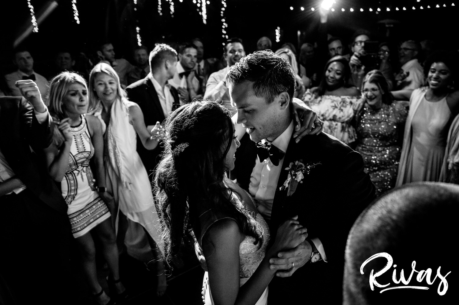 Saddlerock Ranch Summer Wedding | Destination Wedding Photographers | Rivas | A black and white, intimate photo of a bride and groom wrapped up in each others arms dancing while their friends and family encircle them to cheer them on during their wedding reception at Saddlerock Ranch in Malibu. 