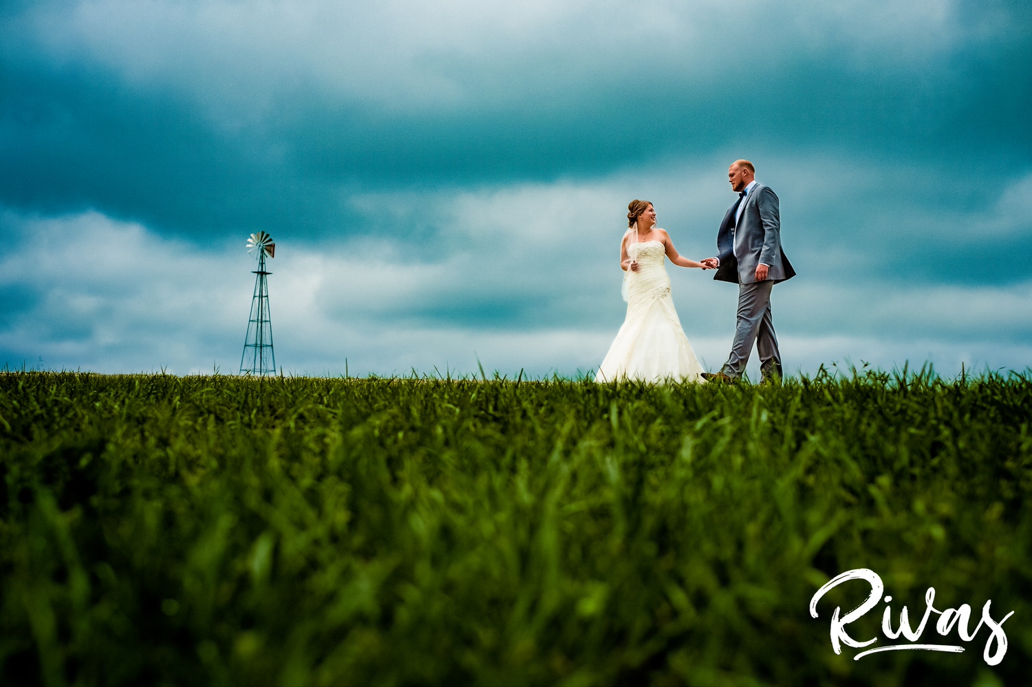 Summer Farm Wedding Sneak Peek | Kansas City Wedding Photographers | A wide photo of a bride and groom holding hands and walking across the horizon line with a single windmill and storm clouds in the background. 