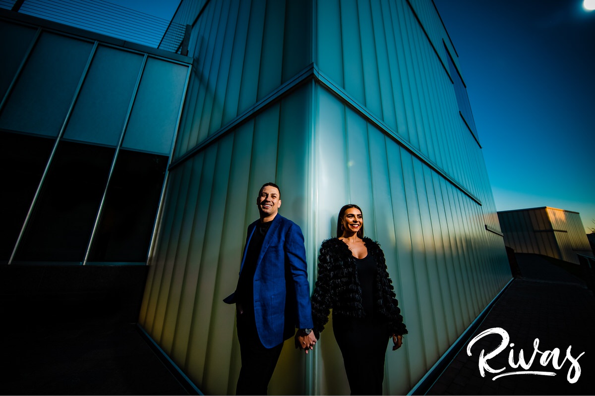 Nelson Atkins Fall Engagement Sneak Peek | Rivas Photography | A dramatic portrait of an engaged couple dressed in black and blue standing against a corner of the Bloch Building at Kansas City's Nelson Atkins Museum of Art holding hands during their sunset engagement session. 