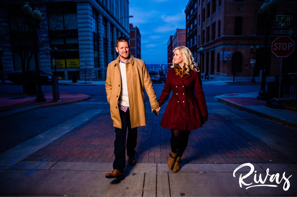 KC Winter Lights Engagement Pictures | Kansas City engagement Pictures | Rivas | A picture of an engaged couple bundled up in dressy winter coats, holding hands and walking down a brick street in downtown Kansas City during their late December Engagement Session. 