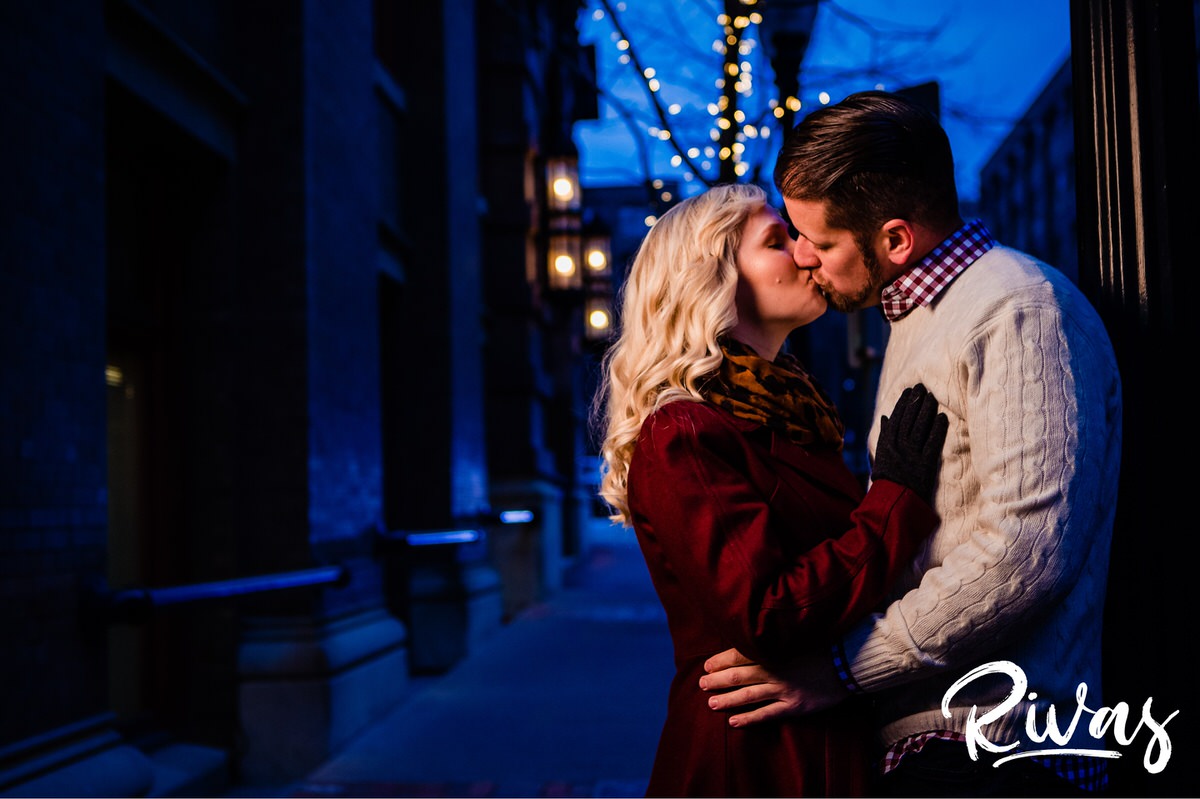 KC Winter Lights Engagement Pictures | Kansas City engagement Pictures | Rivas | A picture taken through a strand of white Christmas lights of an engaged couple leaning up against a light pole, sharing a kiss during their winter engagement session in Kansas City. 