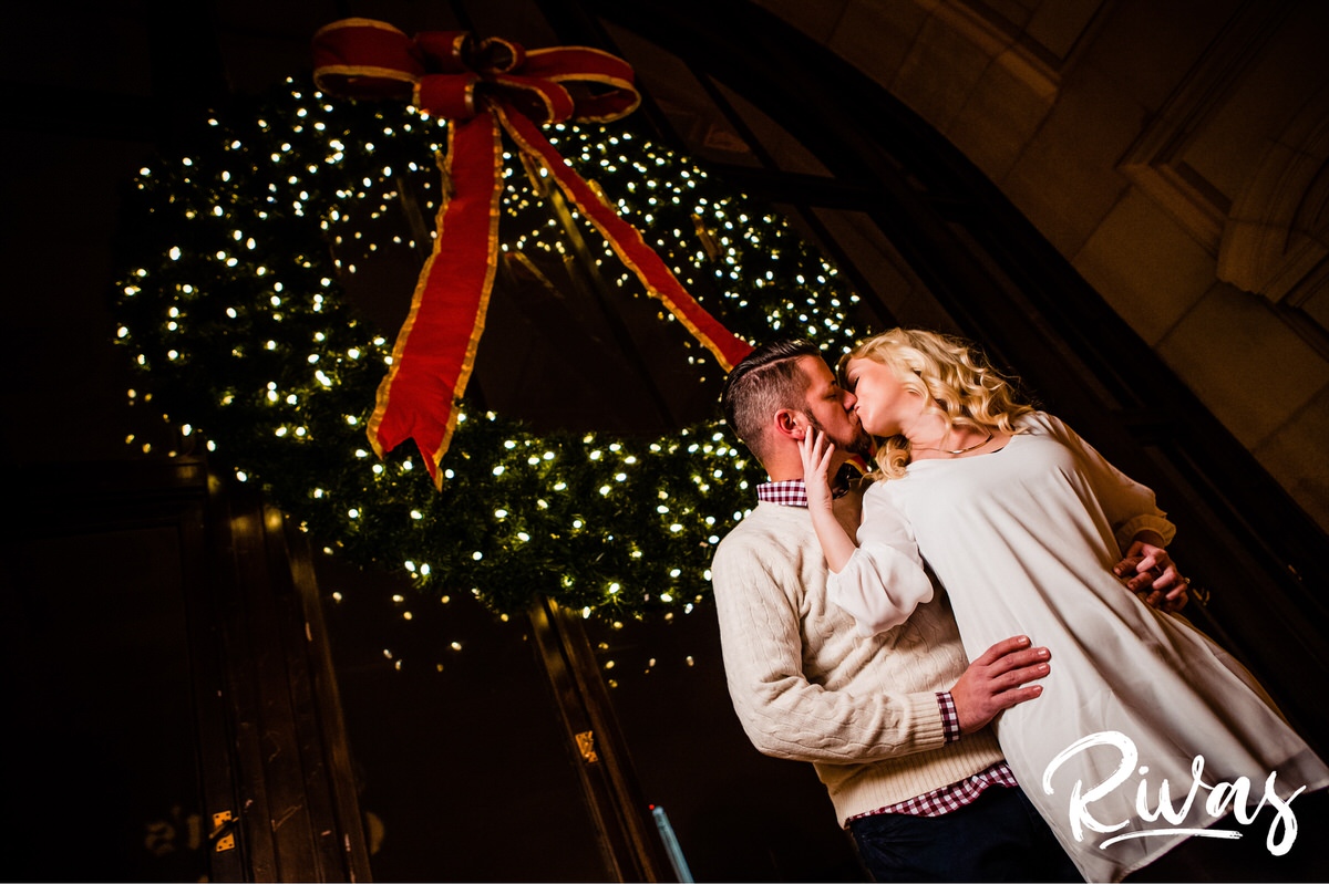 KC Winter Lights Engagement Pictures | Kansas City engagement Pictures | Rivas | A photo taken from the ground up of an engaged couple sharing a kiss as they stand in front of a giant wreath, hanging in a window at Kansas City's Union Station. 