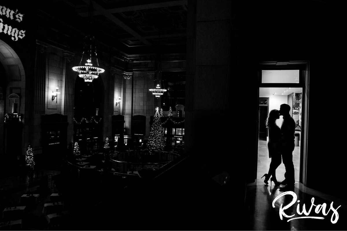 KC Winter Lights Engagement Pictures | Kansas City engagement Pictures | Rivas | A black and white picture with a silhouette of a couple sharing a kiss as they stand in a doorway, with Union Stations Grand Hall lit up behind them during their winter engagement session in Kansas City. 