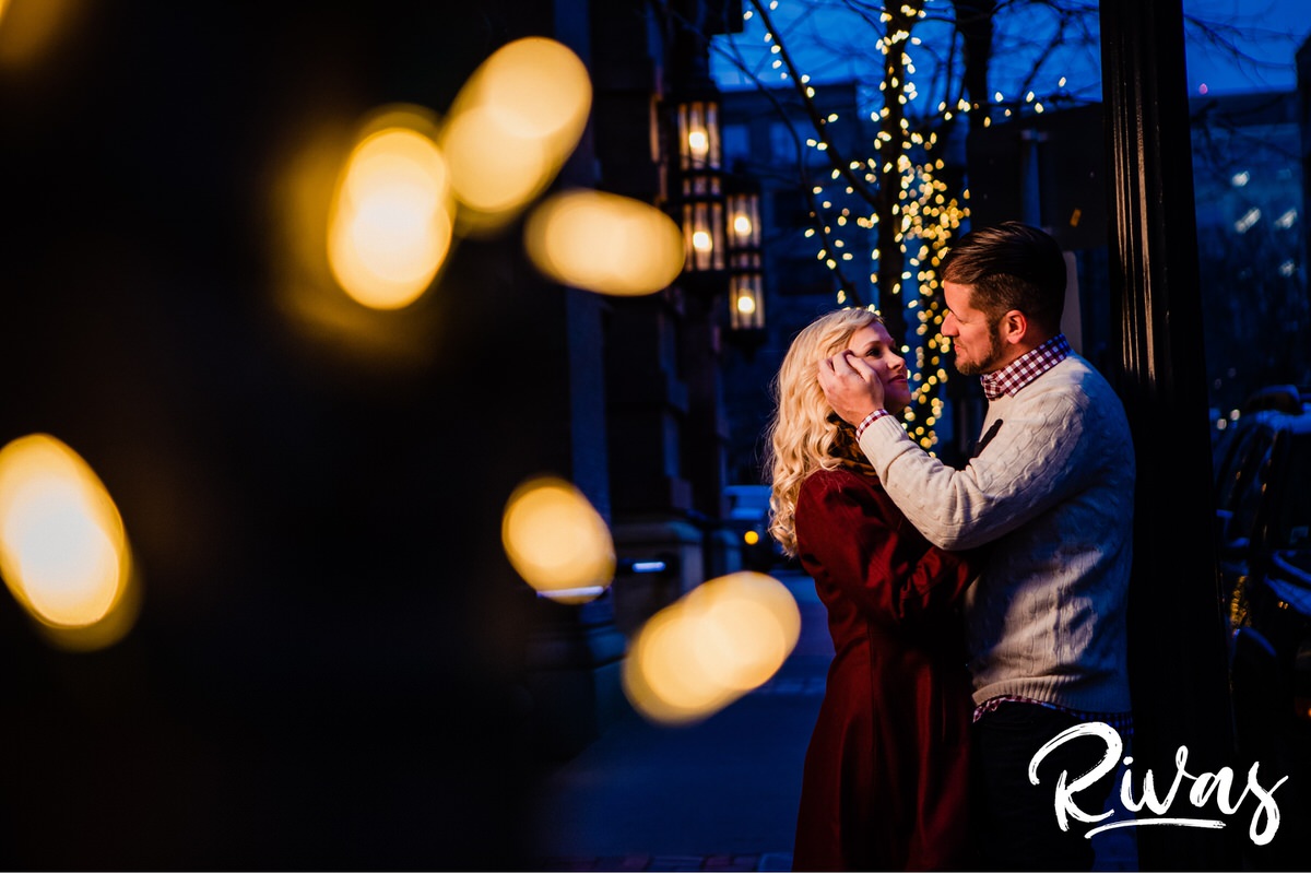 KC Winter Lights Engagement Pictures | Kansas City engagement Pictures | Rivas | A picture taken through a strand of white Christmas lights of an engaged couple leaning up against a light pole, sharing an embrace as he brushes her hair out of her face during their winter engagement session in Kansas City. 