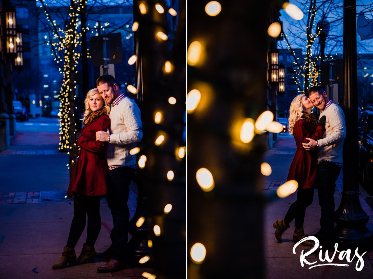 KC Winter Lights Engagement Pictures | Kansas City engagement Pictures | Rivas | A picture taken through a strand of white Christmas lights of an engaged couple leaning up against a light pole, sharing an embrace as he brushes her hair out of her face during their winter engagement session in Kansas City. 
