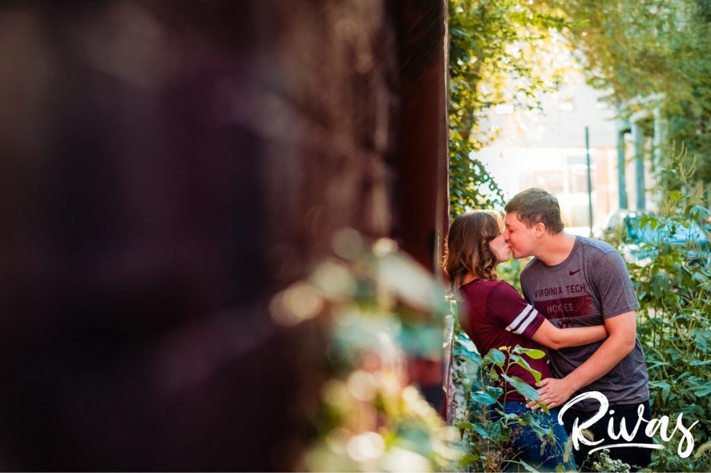 West Bottoms Engagement Pictures | A candid photo of an engaged couple embracing and sharing a kiss taken through a bush. 