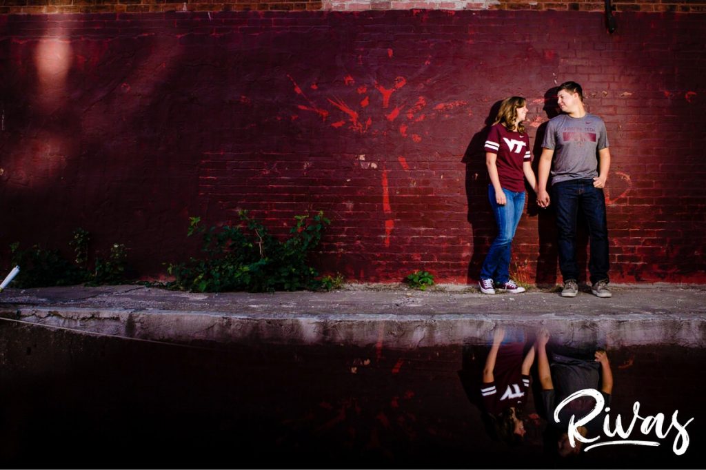 West Bottoms Engagement Pictures | A picture of an engaged couple in Virginia Tech gear standing up against a red brick wall with their reflection in a puddle of water. 