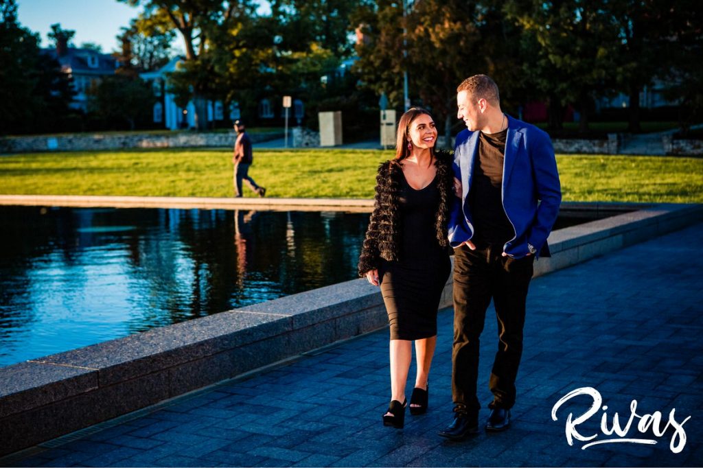 A candid picture of an engaged couple dressed up for date night walking across the driveway by the reflecting pool at the Nelson Atkins Museum of Art during their Colorful Nelson Engagement Session. 