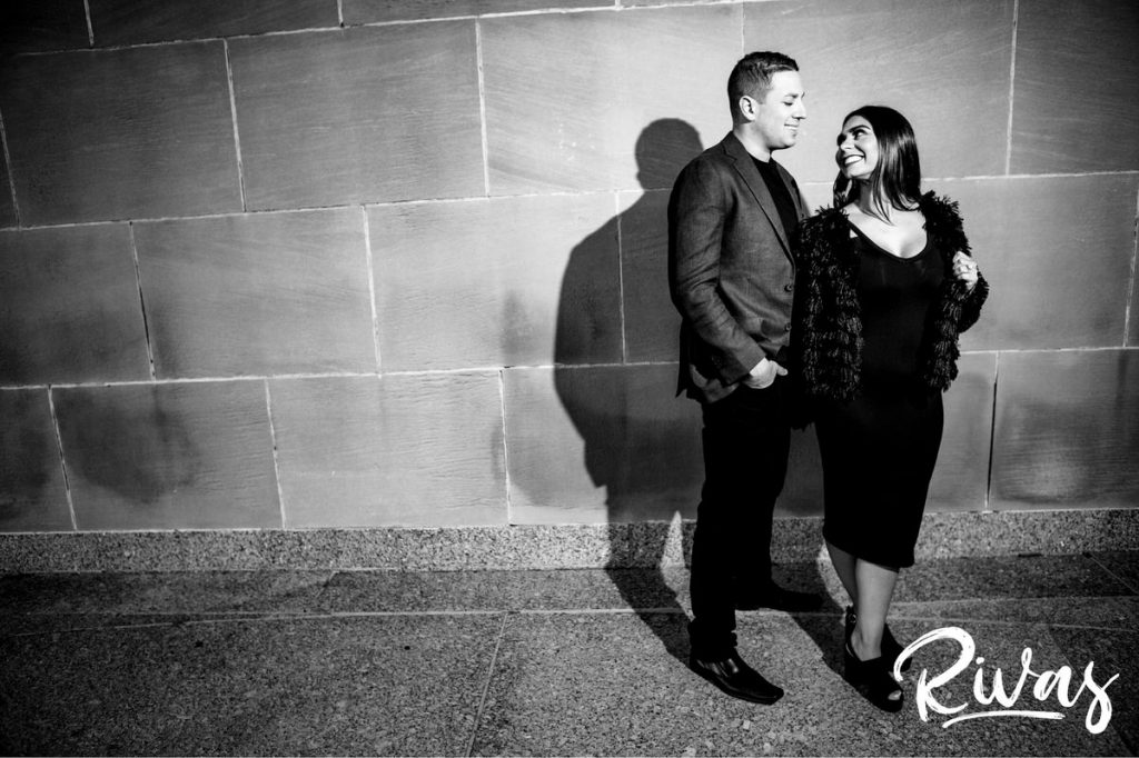 A full length black and white picture of an engaged couple holding hands and looking at each other as they stand in front of a wall during their colorful nelson engagement session in Kansas City. 