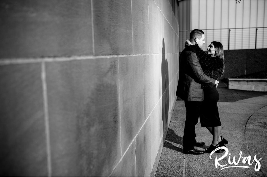 A full length black and white picture of an engaged couple holding hands and looking at each other as they stand in front of a wall during their colorful nelson engagement session in Kansas City. 
