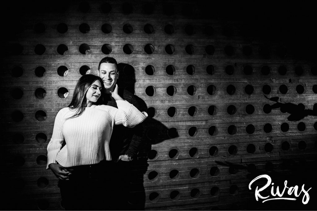 A black and white picture of an engaged couple sharing an embrace as they stand up against a cement wall filled with circles during their colorful Nelson Engagement Session. 