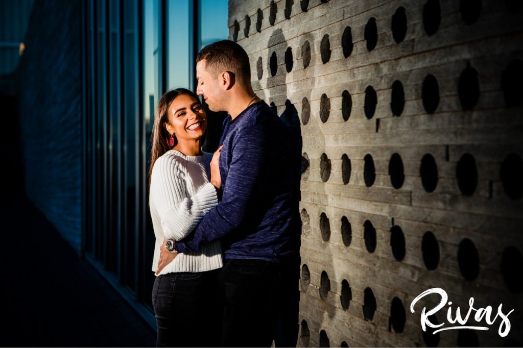 A candid, colorful picture of an engaged couple embracing and laughing together as they stand up against a cement wall filled with circular holes during their colorful Nelson engagement session. 