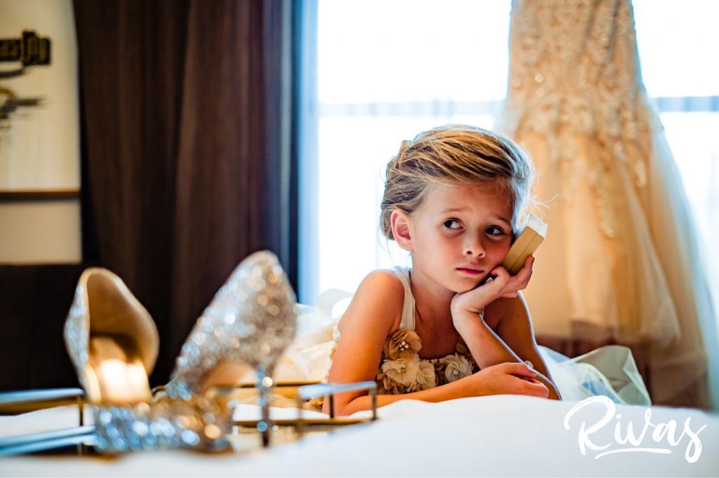 Art Deco Inspired Wedding in Kansas City | A candid picture of a flowergirl sadly holding a box in her hands as she leans on a hotel room bed, in front of a wedding dress hanging in the window. 