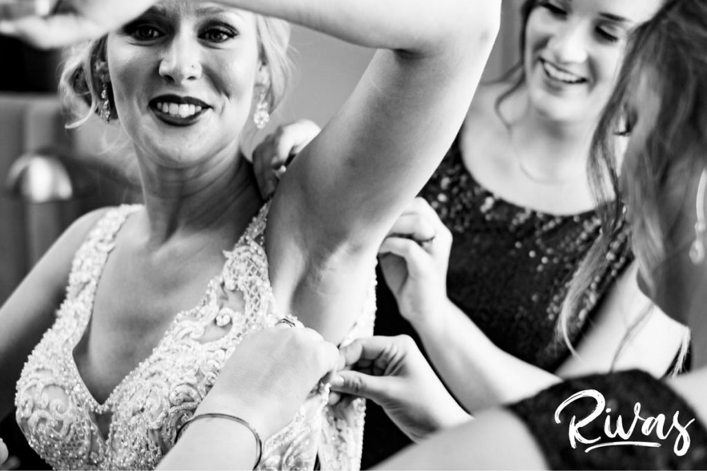 Art Deco Inspired Wedding in Kansas City | A candid black and white picture of a bride holding her arm above her head and laughing as her bridesmaids zip up her wedding dress. 