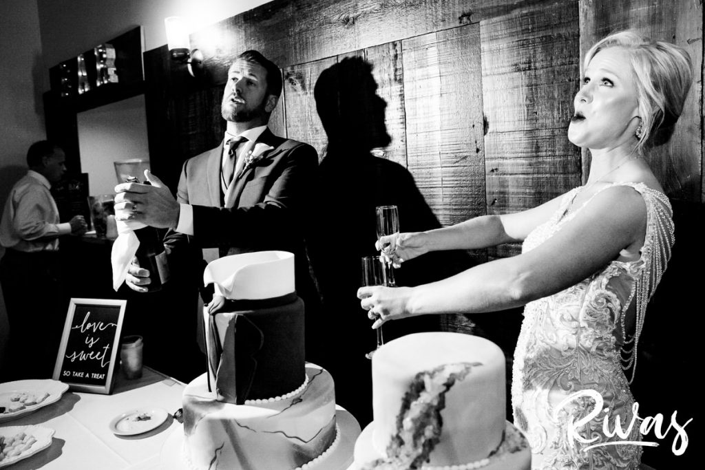 Art Deco Inspired Wedding in Kansas City- A candid black and white picture of a groom popping open a bottle of champagne as his bride cheers him on with champagne flutes in hand. 