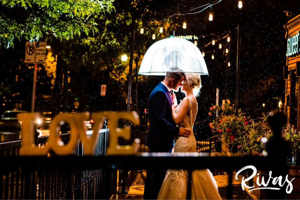 Art Deco Inspired Wedding in Kansas City-A dramatic, nighttime portrait of a bride and groom sharing a kiss as they stand under a clear umbrella on their wedding day. 