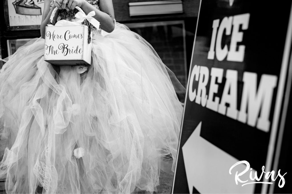 A candid, detailed, black and white image of a flower girl holding her flower bucket standing next to a sign with an arrow on it pointing to an ice cream shop. 