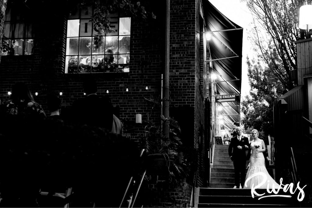 Art Deco Inspired Wedding in Kansas City | A wide, black and white picture of a bride and her father walking down the outside steps next to Californos in Westport, as ice cream shop patrons look on. 
