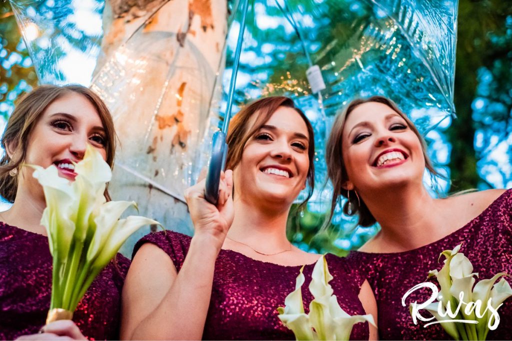 Art Deco Inspired Wedding in Kansas City | A close-up picture of a group of three bridesmaids in red sequined dressed huddling closely under a clear umbrella during a rainy outside wedding ceremony at Kansas City's Californos. 
