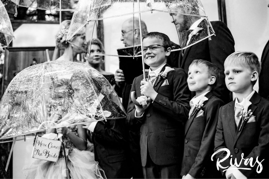 Art Deco Inspired Wedding in Kansas City- A candid, close-up picture of a group of 6 children holding clear umbrellas as they stand with their parents during their outside wedding ceremony at Californos in the rain. 