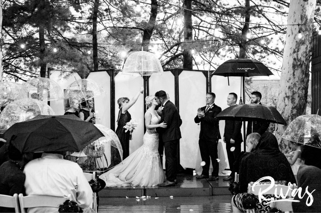Art Deco Inspired Wedding in Kansas City | A wide picture of a bride and groom sharing a kiss at the end of their wedding ceremony as their wedding party holds umbrellas over their heads at Californos. 