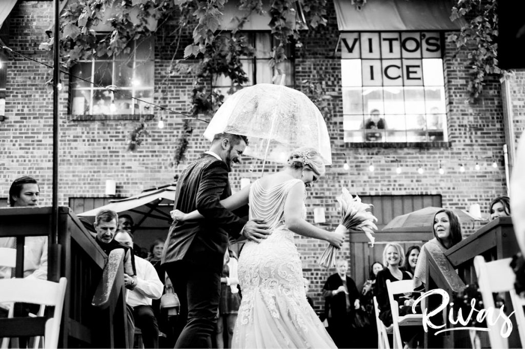 Art Deco Inspired Wedding in Kansas City- A candid black and white picture of a bride and groom sharing an embrace as they walk back down the aisle in the rain after their outside ceremony at Californos. 